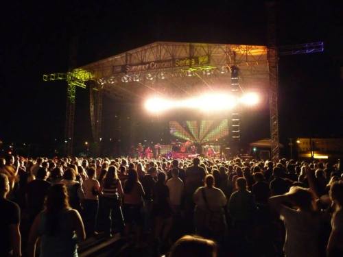 A large crowd enjoys a summer concert at night, with a brightly lit stage and colorful lights in the background.