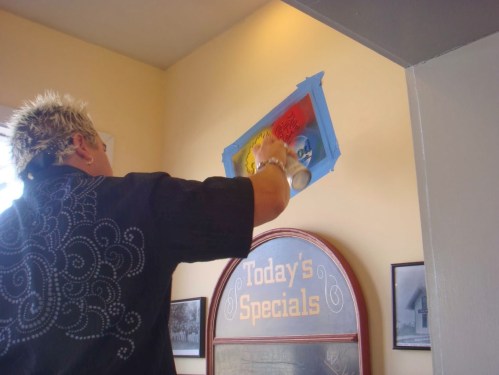 A person with spiky hair spray paints a sign on a wall, with blue tape outlining the area. "Today's Specials" is visible below.
