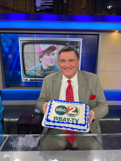 A man in a gray suit holds a cake celebrating WBAY TV's 40th anniversary, with a vintage TV image in the background.
