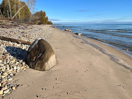 A sandy beach with smooth stones, a large boulder, and calm water under a clear blue sky.