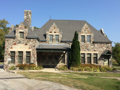 Historic stone building with a gabled roof, labeled "Wisconsin's First Hatchery," surrounded by greenery and blue sky.