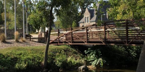 A wooden bridge crosses a small stream, surrounded by trees and a stone building in the background.