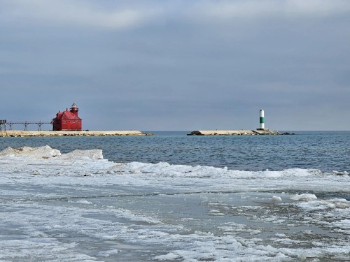 A red lighthouse stands on the shore, with a green and white lighthouse in the distance, surrounded by icy water.
