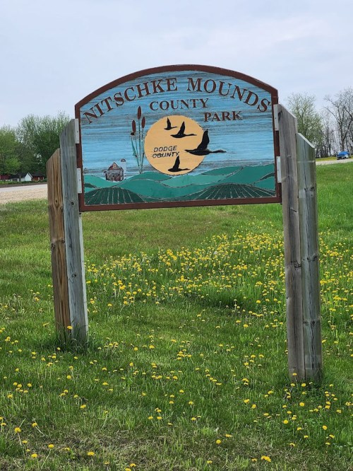 Sign for Nitschke Mounds County Park, featuring a scenic design with fields, birds, and a sunset. Dandelions in foreground.