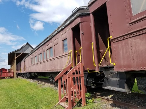 A vintage train car with a maroon exterior, yellow handrails, and a wooden staircase, set against a blue sky.