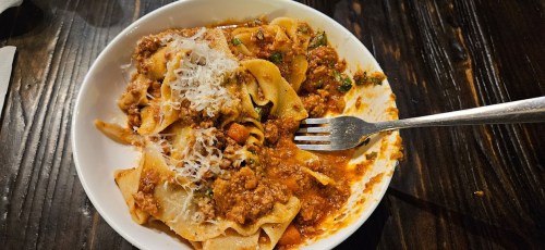 A bowl of pasta with meat sauce, topped with grated cheese, and a fork resting on the side, on a wooden table.