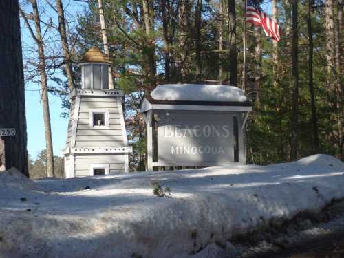 Sign for Beacons Minocqua with a lighthouse structure beside it, surrounded by snow and trees, and an American flag.