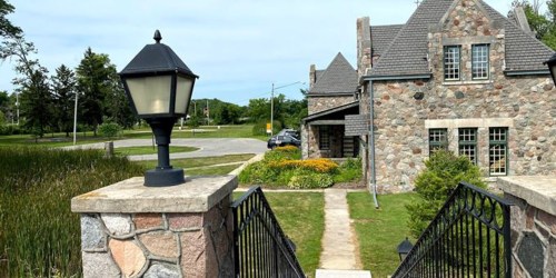 A stone house with a gray roof, surrounded by greenery and flowers, viewed from a pathway with a lamp post.