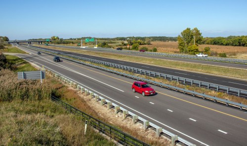 A red SUV drives on a highway with sparse traffic, surrounded by fields and trees under a clear blue sky.