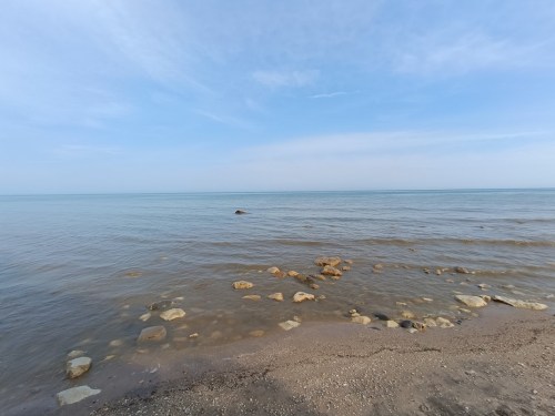 A calm shoreline with smooth stones, gentle waves, and a clear blue sky over a tranquil body of water.