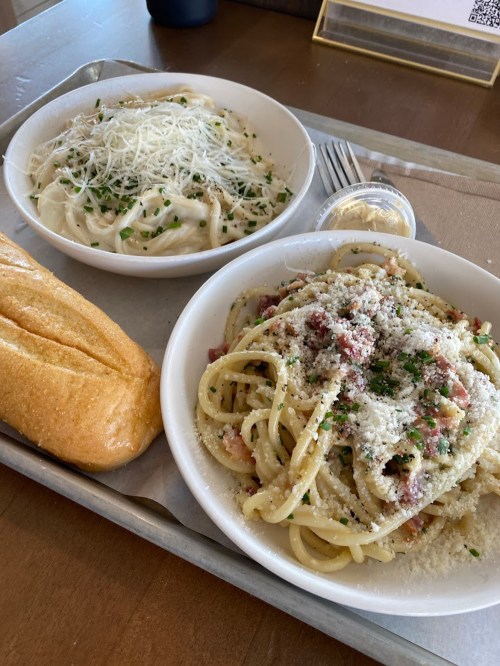 Two bowls of pasta on a tray: one with creamy sauce and cheese, the other with bacon and herbs, alongside a bread roll.