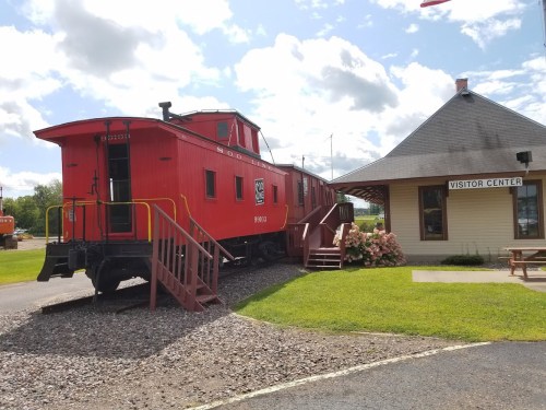 A red caboose next to a visitor center, surrounded by green grass and flowers under a partly cloudy sky.