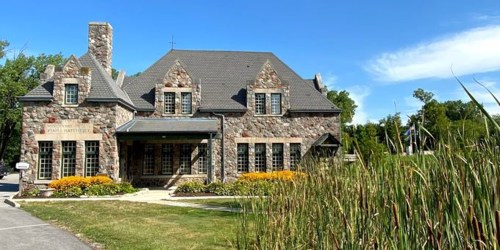 A stone building with a gray roof, surrounded by greenery and colorful flowers under a clear blue sky.