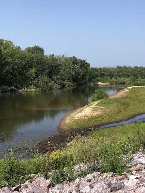 A serene river scene with lush greenery and a sandy bank under a clear blue sky.