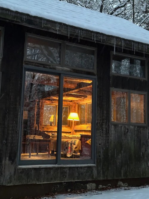 A cozy, warmly lit interior visible through a snowy window, surrounded by trees and icicles.