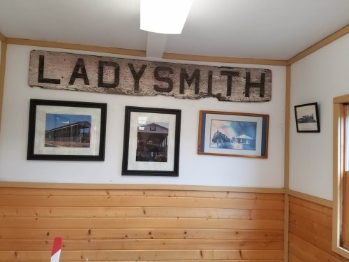 A wooden sign reading "LADYSMITH" hangs on a wall with framed photos in a room with wooden paneling.