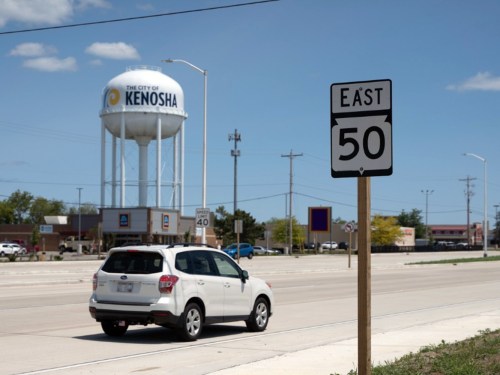 A car drives past a road sign for East 50, with a Kenosha water tower in the background under a clear blue sky.