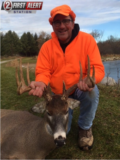 A man in an orange jacket poses with a deer, showcasing its antlers near a pond and trees.