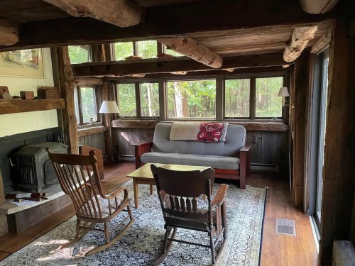 Cozy living room with wooden beams, a gray sofa, rocking chairs, and large windows overlooking a forested area.