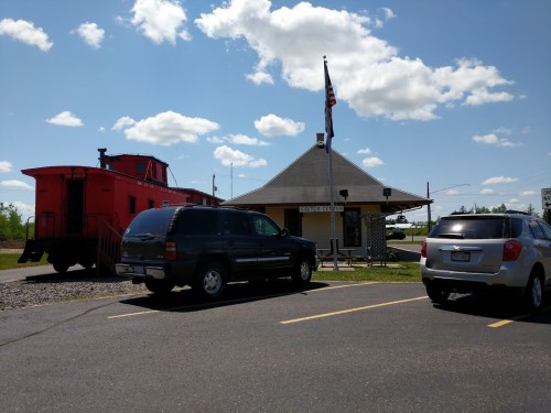 A historic train depot with a red caboose, surrounded by parked cars and a blue sky with fluffy clouds.
