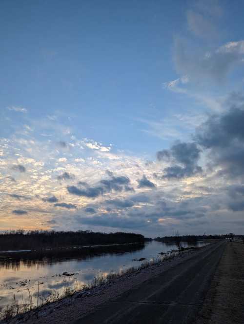 A serene river scene at dusk, with a cloudy sky reflecting on the water and a path along the riverbank.