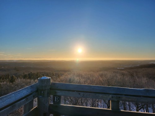 A scenic view from a wooden lookout, featuring a bright sun rising over a misty landscape and distant hills.