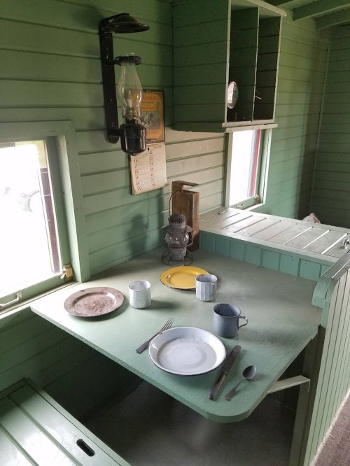 Interior of a green-painted cabin with a table set for a meal, featuring plates, cups, and a lantern on the wall.
