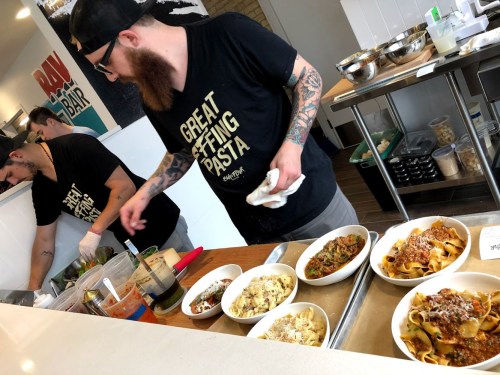 Two chefs in black shirts prepare pasta dishes in a kitchen, with various plates of pasta displayed on a counter.