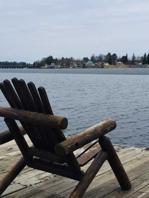 A wooden chair on a dock overlooking a calm lake with a distant shoreline and cloudy sky.