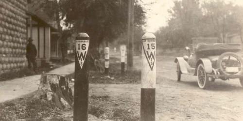 Historic black and white photo of road markers and an early automobile near a rustic building.