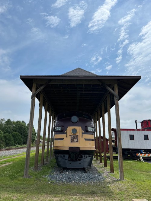 A vintage train displayed under a wooden shelter, with a blue sky and clouds in the background.