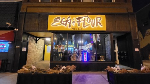 Exterior view of Egg & Flour Pasta Bar, featuring a bright sign and an inviting atmosphere with open doors.