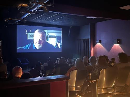 A dimly lit theater with an audience watching a film on a large screen, featuring a close-up of a man's face.