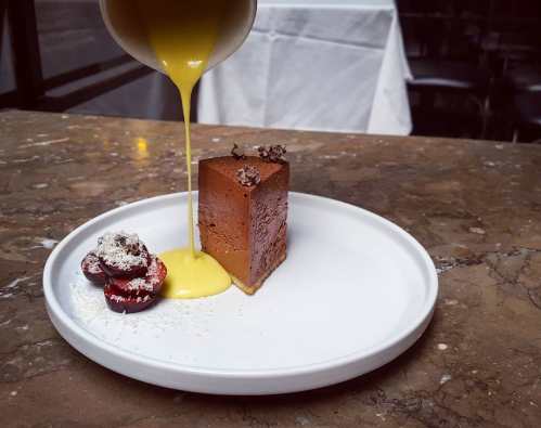 A slice of chocolate dessert on a white plate, topped with cherries and drizzled with yellow sauce.