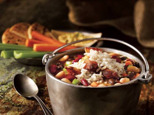 A pot of hearty bean soup topped with rice, served with celery, carrots, and bread on a rustic table.