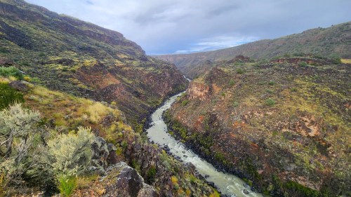 A winding river flows through a rugged canyon surrounded by green hills and rocky cliffs under a cloudy sky.