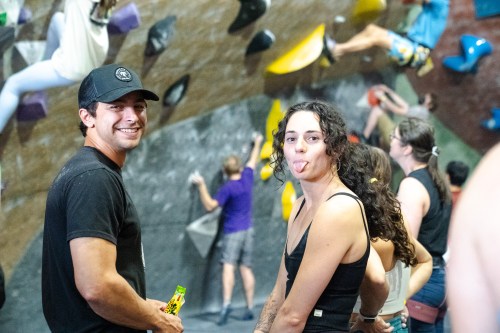 Two climbers smile at the camera in a bouldering gym, with others climbing in the background. One makes a playful face.