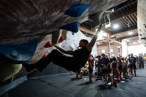 A climber hangs from a bouldering wall while a crowd watches in a climbing gym.