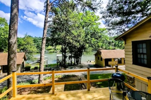 Scenic view of a lake surrounded by trees, with cabins and a deck in the foreground under a bright blue sky.