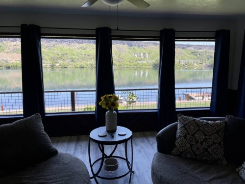 Cozy living room with two sofas, a table, and large windows overlooking a serene river and green hills.