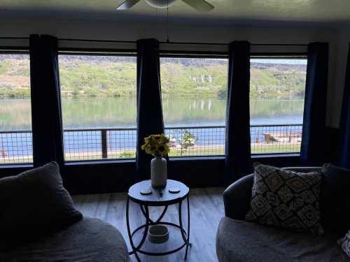 Cozy living room with two sofas, a table, and large windows overlooking a serene river and green hills.