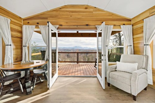 Cozy interior with wooden walls, a round table, a chair, and open doors leading to a scenic outdoor deck.