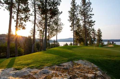 A serene golf course at sunset, surrounded by tall trees and a view of a lake in the background.