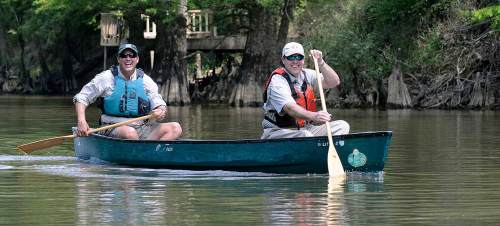 Two men paddle a canoe on a calm river, surrounded by lush greenery and trees.
