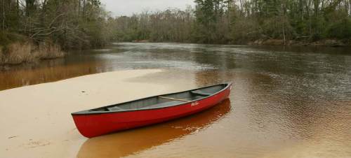 A red canoe rests on a sandy bank by a calm river, surrounded by trees and a cloudy sky.