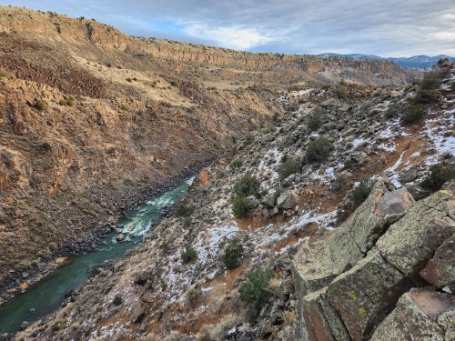 A scenic view of a winding river surrounded by rocky hills and sparse vegetation under a cloudy sky.