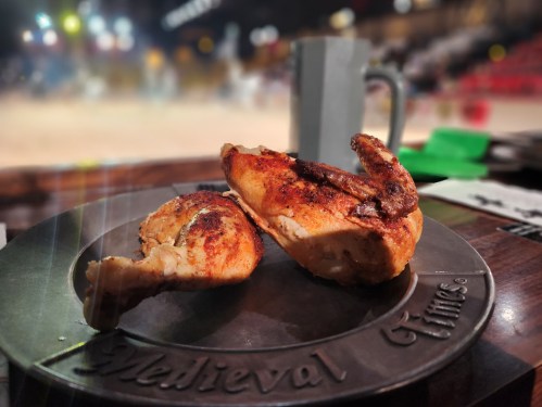 A plate with two pieces of roasted chicken, set against a blurred background of a medieval-themed event.