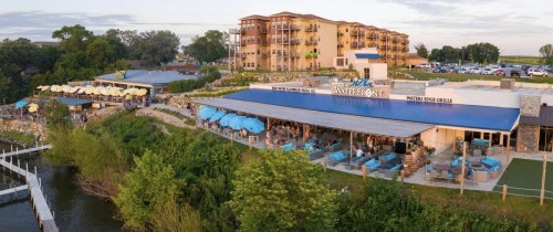 A waterfront restaurant with outdoor seating, surrounded by greenery and a multi-story building in the background.