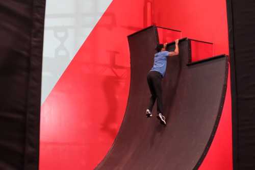 A person climbs a vertical ramp against a red backdrop in a sports or obstacle course setting.