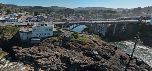 A coastal town with a bridge, rocky shoreline, and buildings nestled among trees under a clear blue sky.